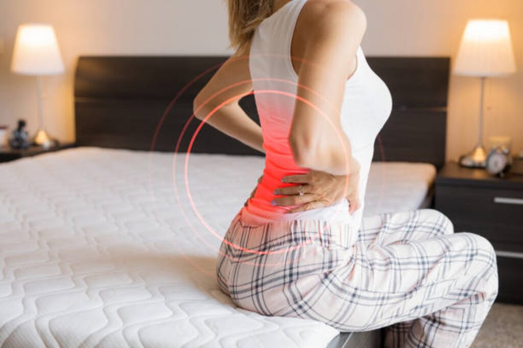 Person kneeling on a bed with hands on lower back, indicating back pain in a bedroom setting with red circular pain marks around the lower back area