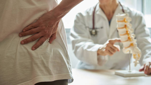 Patient with back pain is examined by a doctor who points to a spine model on the table behind themy.