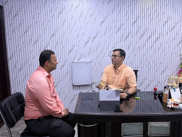 Two men sit across a glass desk in an office, one in a peach shirt talking while the other writes notes.