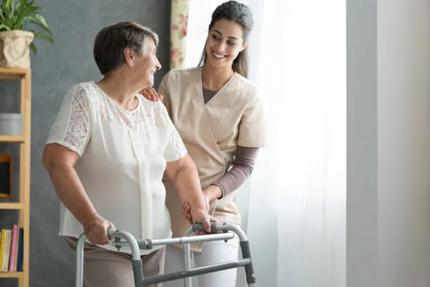 Caregiver helping an elderly woman stand with a walker, both smiling in a bright room.