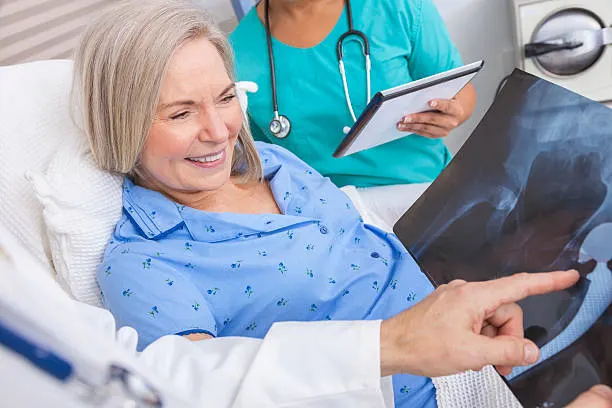 Older woman in a hospital gown lying in bed while a nurse and doctor review an X-ray film, the patient pointing at it.
