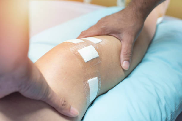 Close-up of a patient's knee with white adhesive bandages taped in place, held by a caregiver.